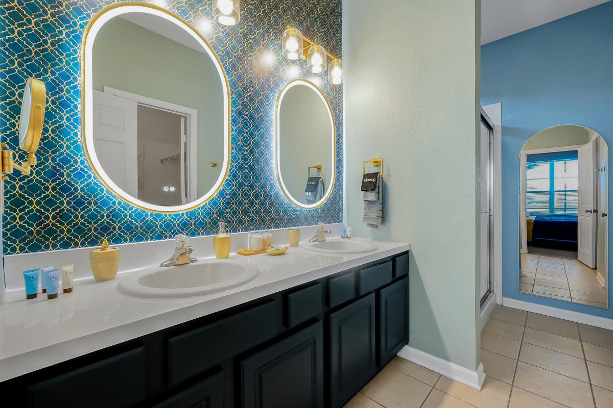 Bright spa-style en-suite with double vanity, backlit mirrors, gold accents, and coastal tones.